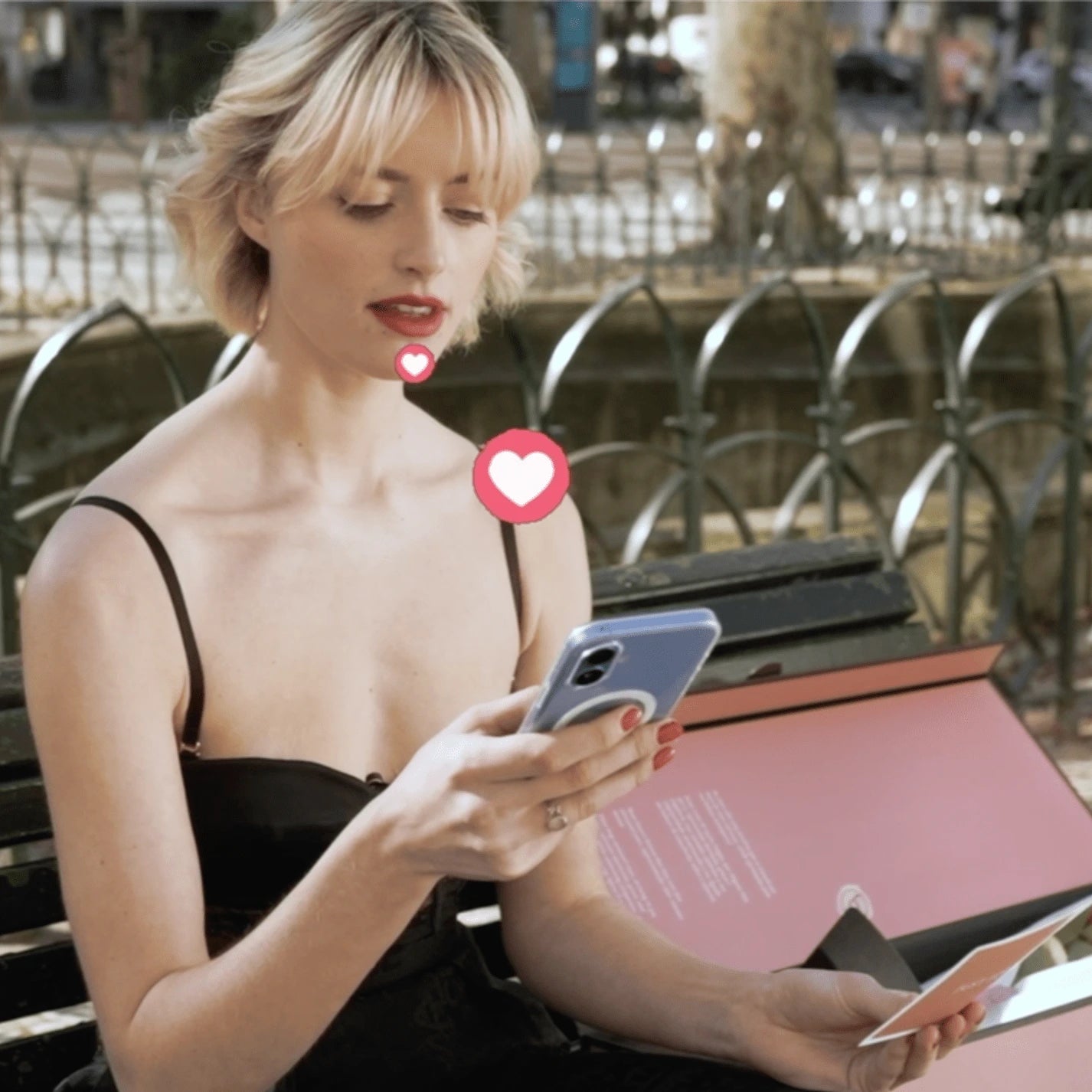 Mulher loira com vestido preto sentada ao ar livre usando smartphone, com caixa de presente rosa da Q_Roses ao lado. Um ícone de coração rosa flutua digitalmente sobre a imagem, ilustrando a conexão entre tecnologia e emoção. Ao fundo, ambiente urbano com arquitetura moderna desfocada.